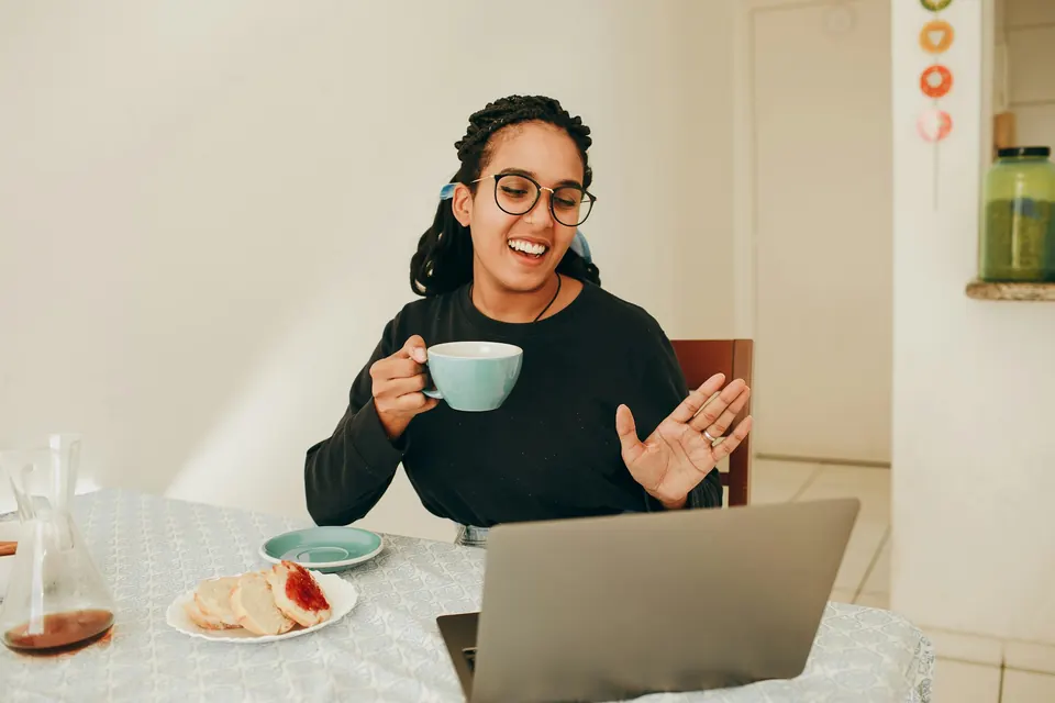 A woman smiling and holding a mug of tea during a video call on her laptop