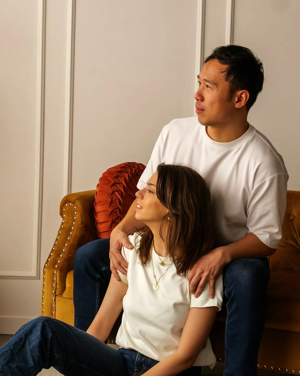A couple relaxing together at home, one partner seated on an orange sofa and the other leaning back against them